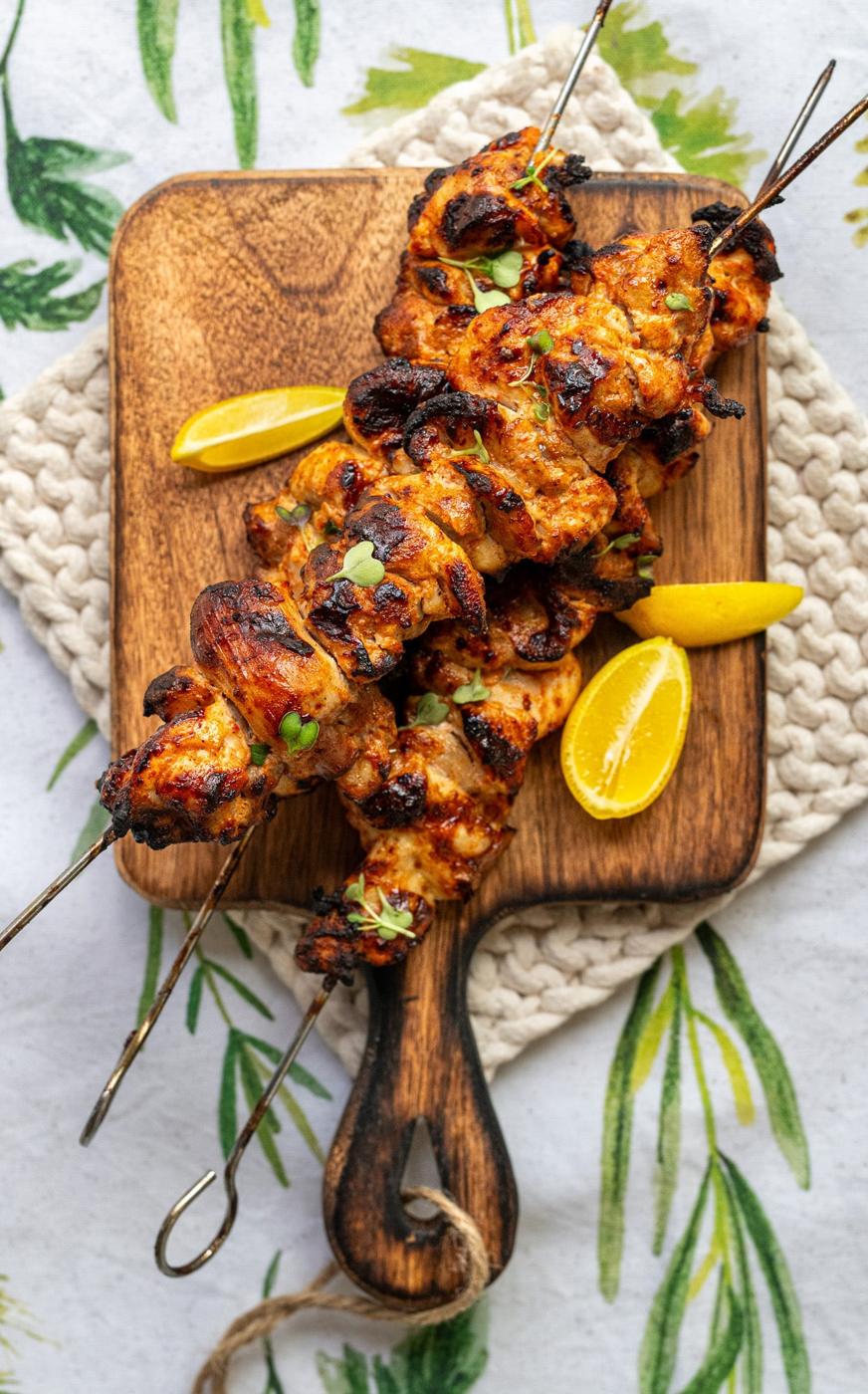 Grilled Chicken Kebabs with spices served on a wooden board, garnished with fresh herbs and accompanied by lemon wedges. The board rests on a textured cloth with a leafy pattern.