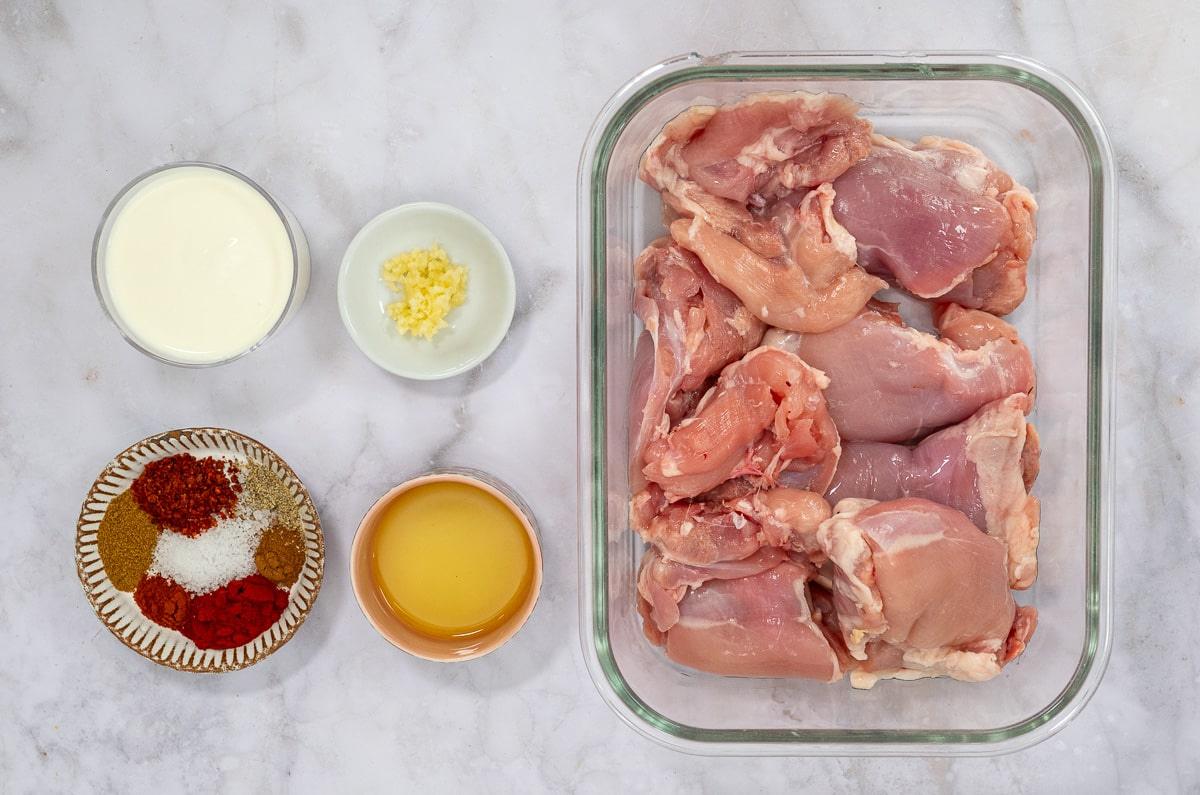 Raw chicken thighs in a glass container, surrounded by small bowls of yogurt, minced garlic, spices (including paprika and cumin), and oil—perfect for marinating flavorful Chicken Kebabs—all arranged on a white marble surface.