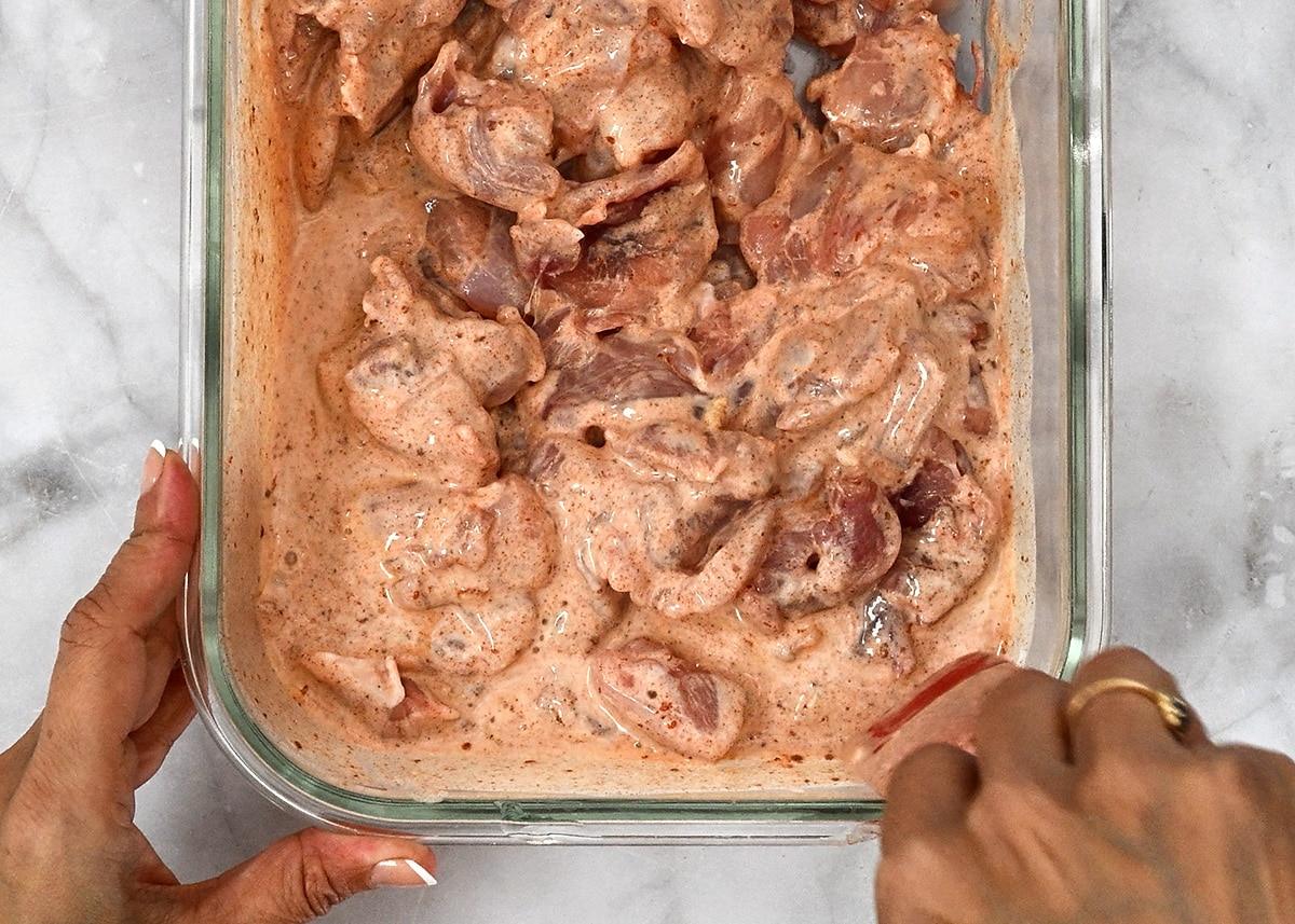 A person stirs raw chicken pieces coated in a creamy, spiced marinade inside a glass dish, preparing flavorful chicken kebabs for cooking. The dish is placed on a marble countertop.