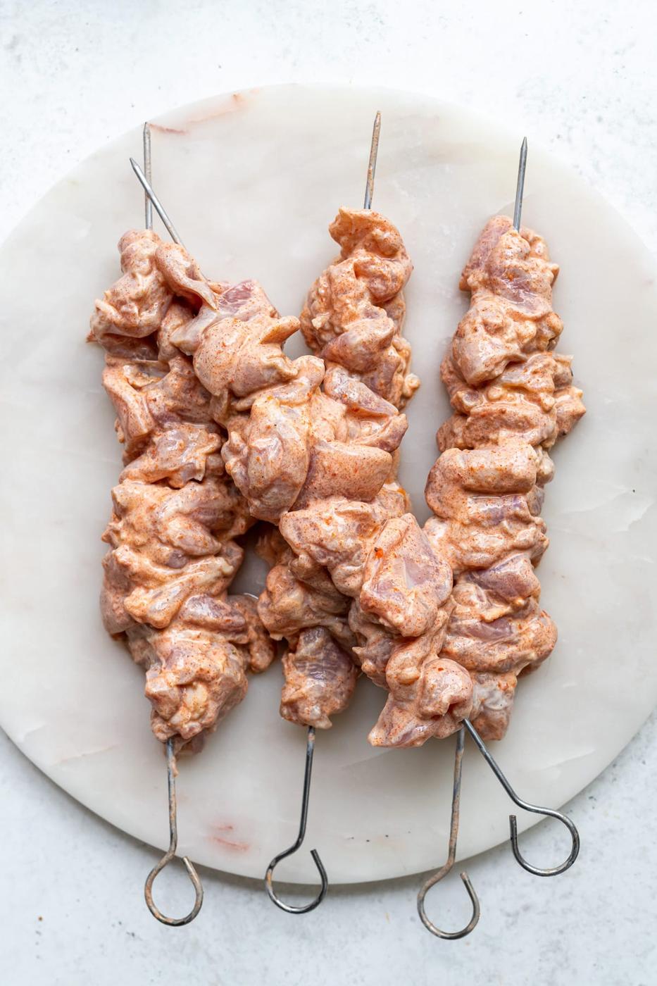 Three metal skewers with marinated raw chicken pieces are arranged on a round white plate, ready to be grilled or cooked. These vibrant Chicken Kebabs are coated in a flavorful reddish-orange marinade.