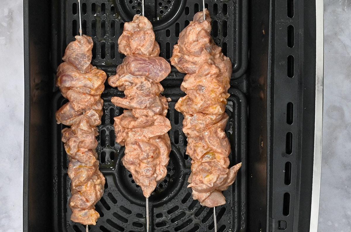 Three skewers of marinated raw chicken kebabs are placed inside an air fryer basket, ready to be cooked. The meat is coated in a light-colored marinade. The air fryer tray is black with a perforated surface.