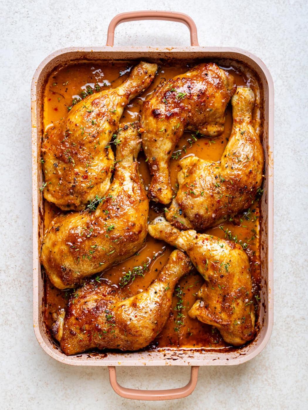 Baked chicken leg quarters with golden, crispy skin and herbs, arranged in a rectangular pan with a rich, seasoned sauce.