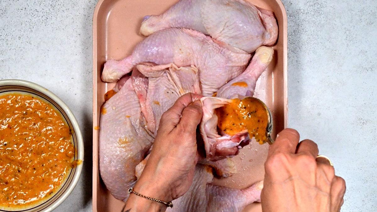 A person stuffs seasoned marinade under the skin of raw chicken quarters on a tray, with a bowl of marinade beside them on a white surface.