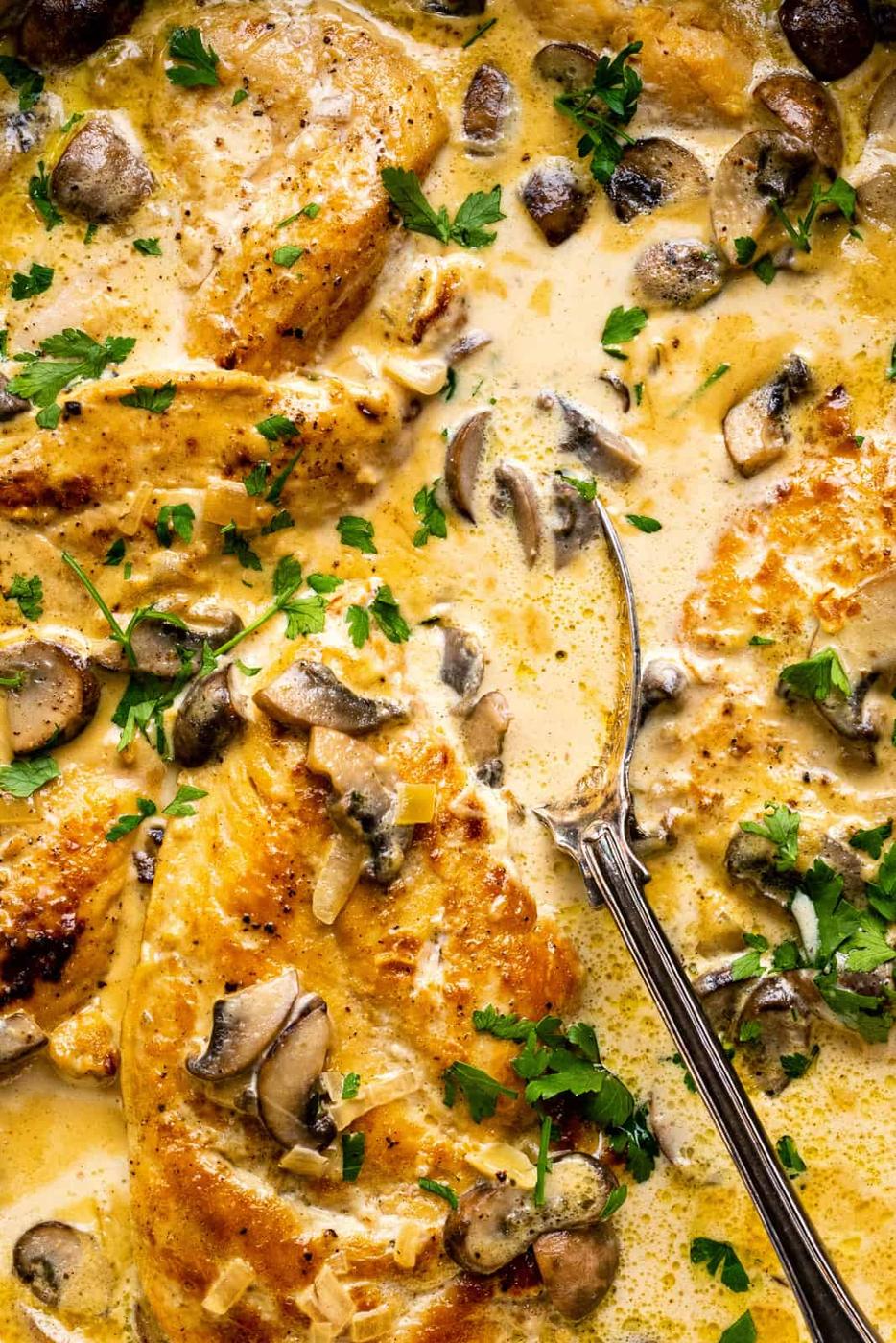 Close-up of creamy Chicken Marsala breasts in a mushroom sauce, garnished with chopped parsley, with a spoon resting in the pan among the cooked mushrooms and chicken.