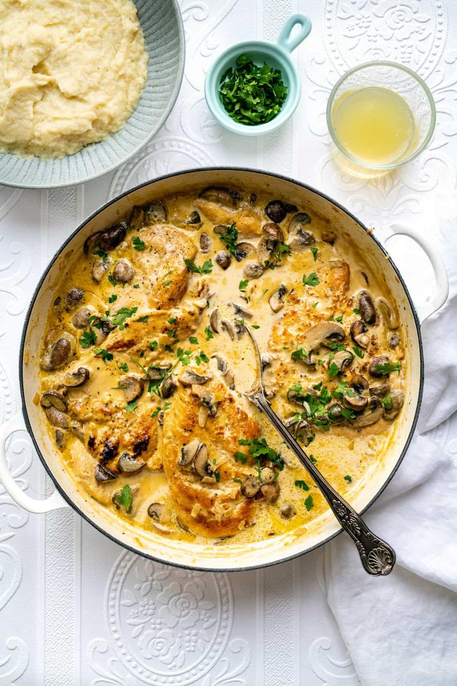 A skillet filled with creamy Chicken Marsala and mushrooms, garnished with chopped herbs. Nearby are a bowl of mashed potatoes, a small bowl of fresh herbs, and a glass of light-colored liquid, all on a white tablecloth.