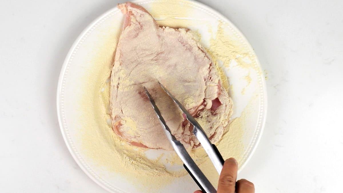A hand uses metal tongs to coat a raw chicken breast with flour on a white plate, preparing it for Chicken Marsala.