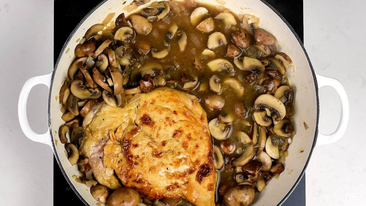A white pot on a stovetop filled with cooked chicken breast and sautéed mushrooms in a rich Chicken Marsala brown sauce. The mushrooms are sliced and spread around the chicken.
