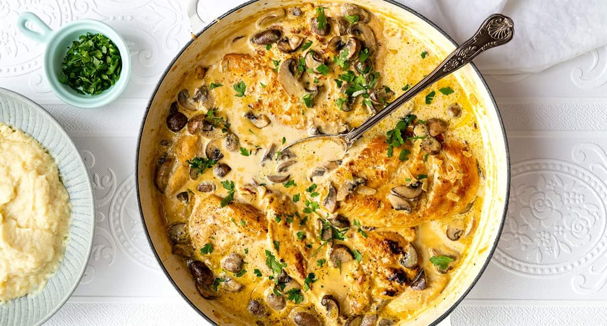 Skillet of creamy Chicken Marsala with mushrooms, topped with chopped parsley, sits beside a bowl of mashed potatoes and a small dish of fresh herbs on a white patterned table.