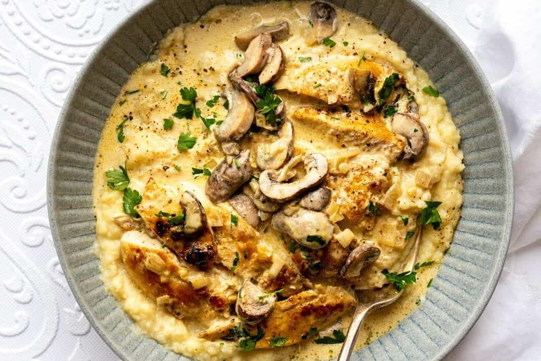 A bowl of creamy polenta topped with Chicken Marsala-style grilled chicken strips, sautéed mushrooms, and chopped herbs. A spoon rests in the bowl. Nearby are a cup of chopped herbs and a glass of light-colored liquid on a white tablecloth.