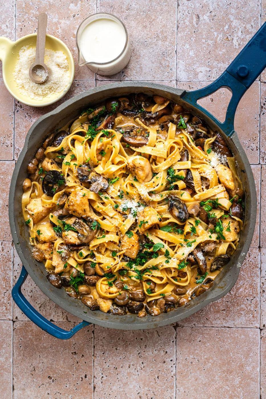 A skillet filled with tagliatelle pasta, mushrooms, and chicken, garnished with chopped parsley. Nearby are a bowl of grated parmesan with a wooden spoon and a small pitcher of cream, all on a tiled surface.