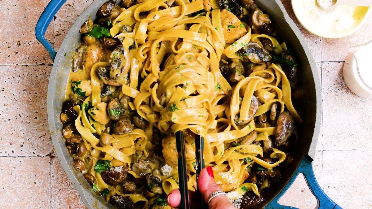 A hand holding tongs lifts creamy fettuccine pasta with mushrooms and parsley from a blue skillet, set on a tiled countertop.