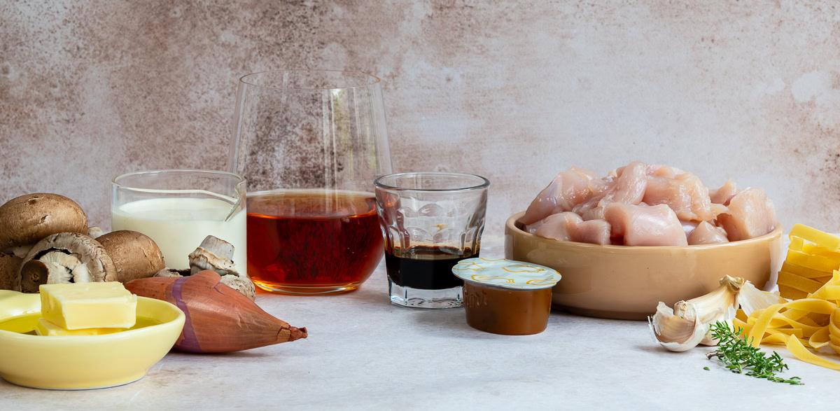A variety of ingredients including raw chicken cubes, uncooked pasta, mushrooms, a shallot, garlic, butter, cream, a glass of wine, soy sauce, a small sauce cup, and fresh herbs arranged on a kitchen counter.