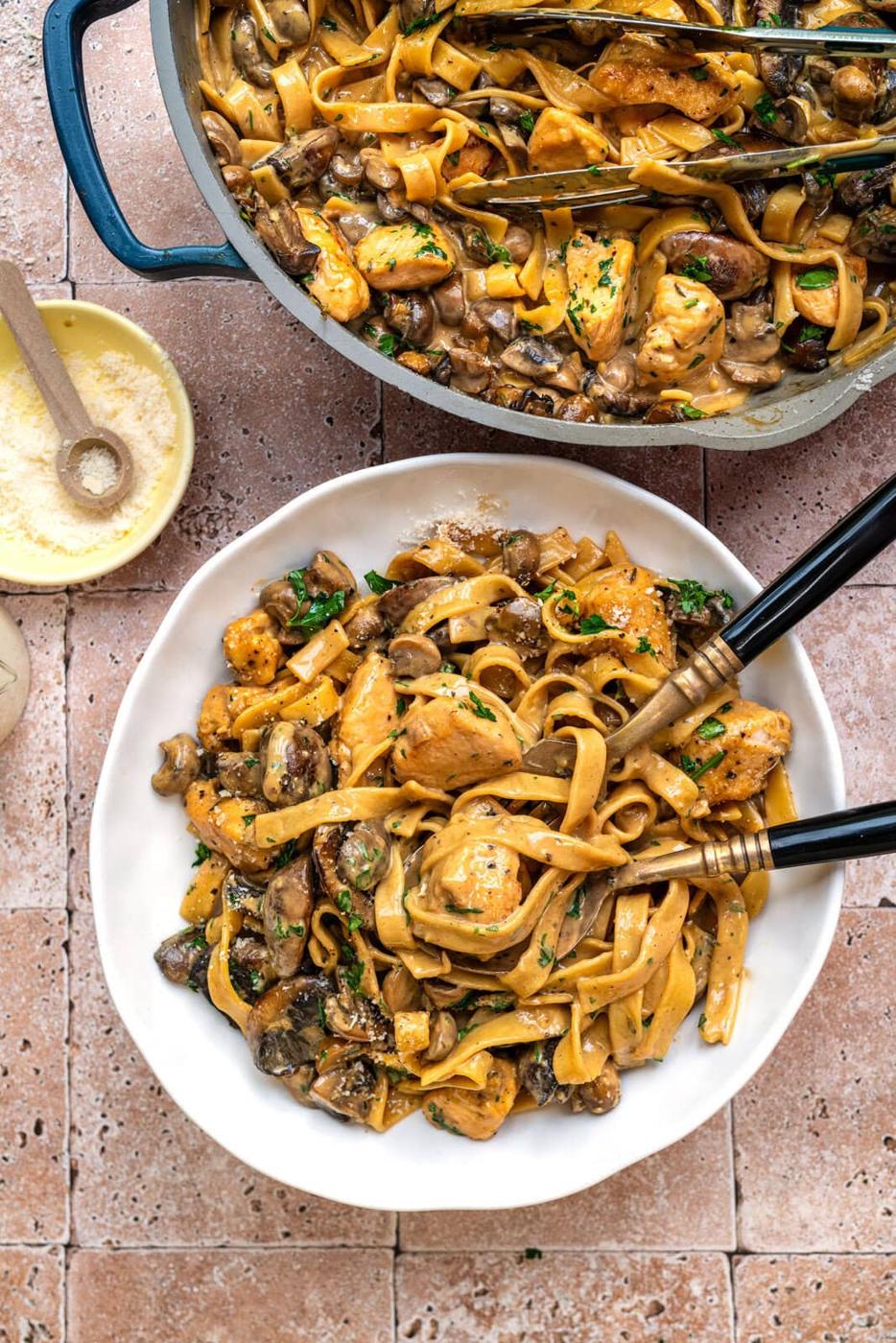 A white plate of fettuccine pasta with chicken, mushrooms, and herbs sits on a tiled surface beside a pot of the same dish. Black-handled cutlery, grated cheese, and a small bowl of salt are also visible.