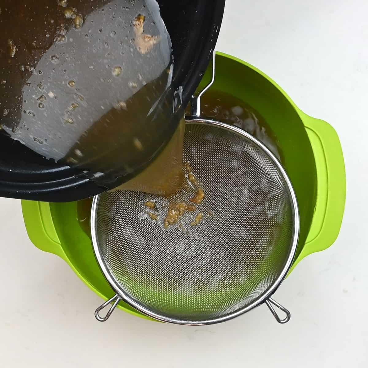 A dark liquid, likely chicken soup from carcass, is being poured from a pot through a metal strainer into a green bowl, separating the solids from the broth.