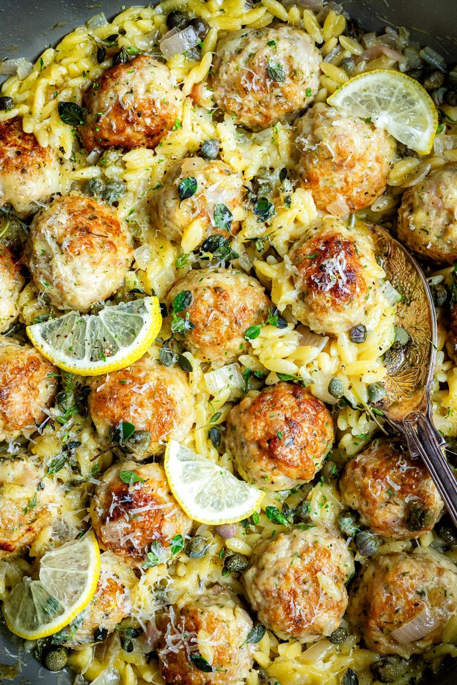 Golden-brown Chicken Piccata Meatballs with orzo pasta, garnished with lemon slices, capers, grated cheese, and fresh herbs, served in a pan with a wooden spoon.
