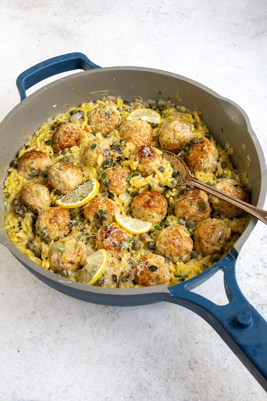 A large blue skillet filled with Chicken Piccata Meatballs, orzo pasta, lemon slices, and herbs, with a serving spoon resting inside. The dish is creamy and garnished with Parmesan cheese.