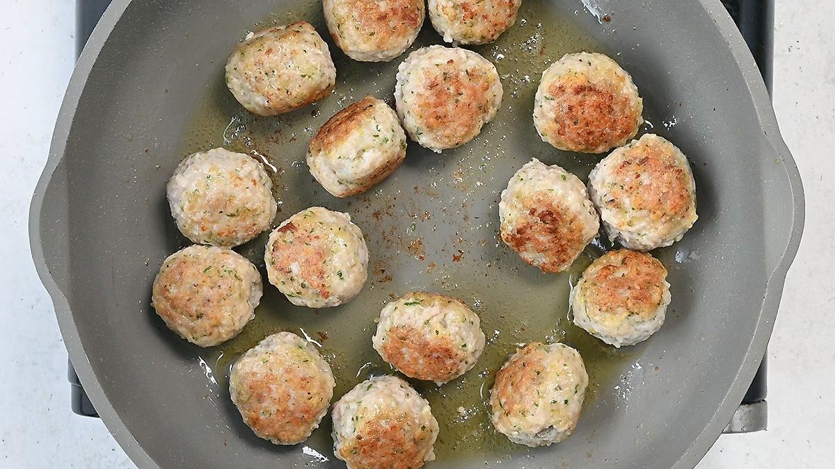 A gray frying pan with several browned Chicken Piccata Meatballs cooking in oil, arranged in a single layer on the pan’s surface.