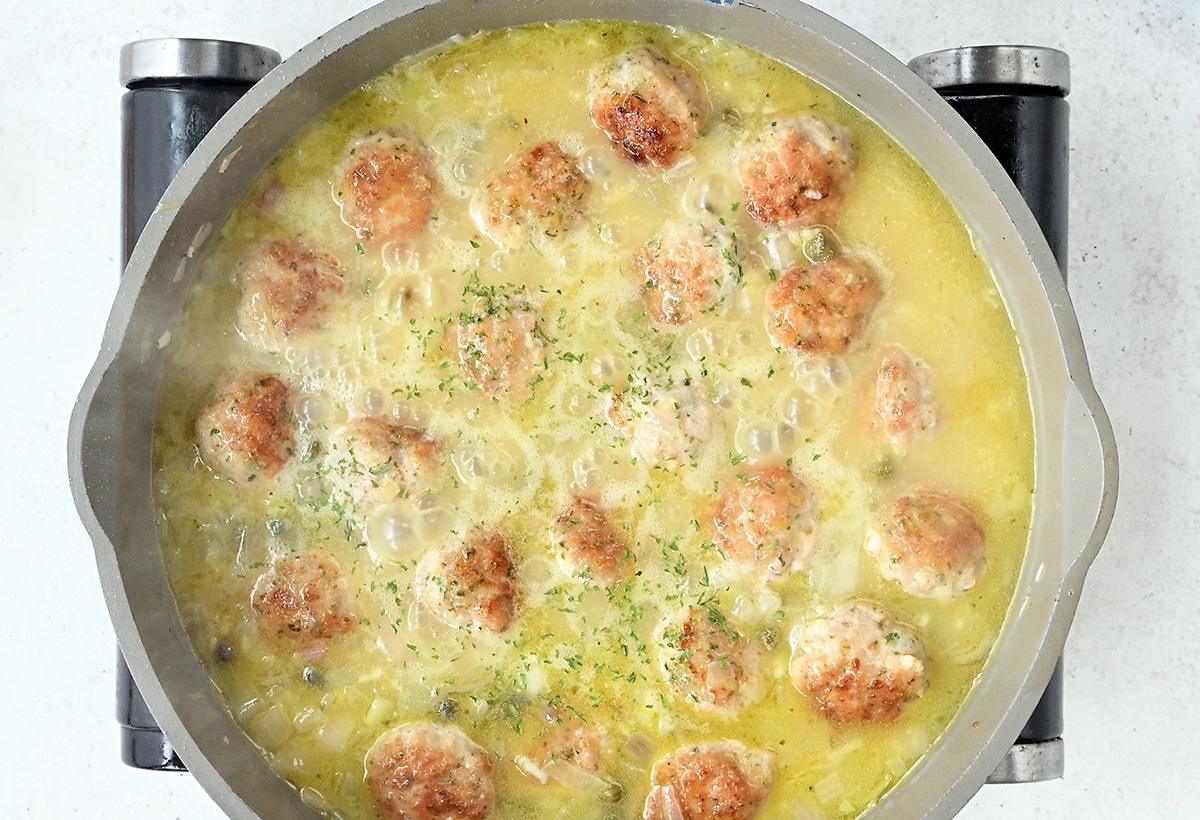 A skillet filled with Chicken Piccata Meatballs simmering in a creamy yellow sauce, garnished with chopped herbs, sits on a stovetop.
