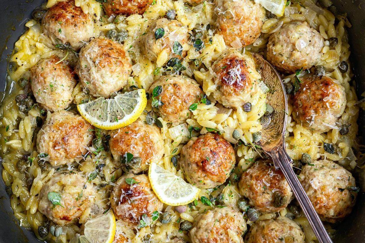 A skillet filled with Chicken Piccata Meatballs on creamy orzo pasta, garnished with lemon slices, grated cheese, capers, and fresh herbs. A spoon rests beside the meatballs.