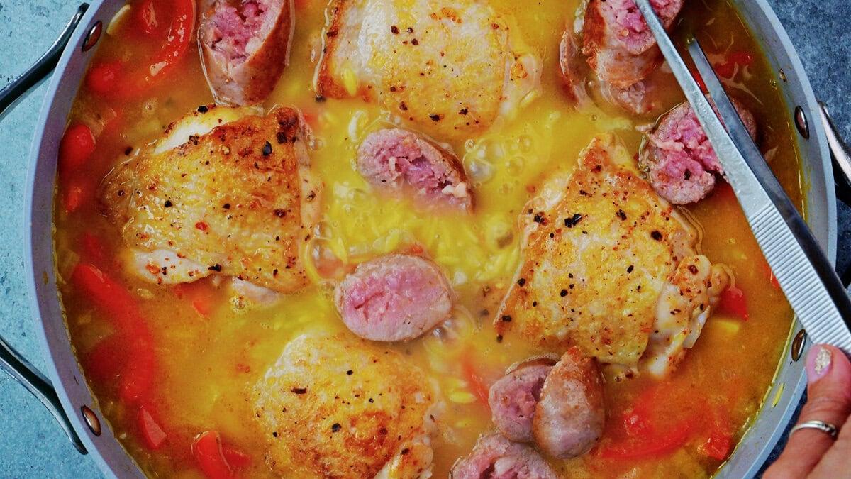 Seared chicken thighs set skin side up on orzo, sliced sausage and red bell peppers in a pan of chicken stock, ready to go into the oven.