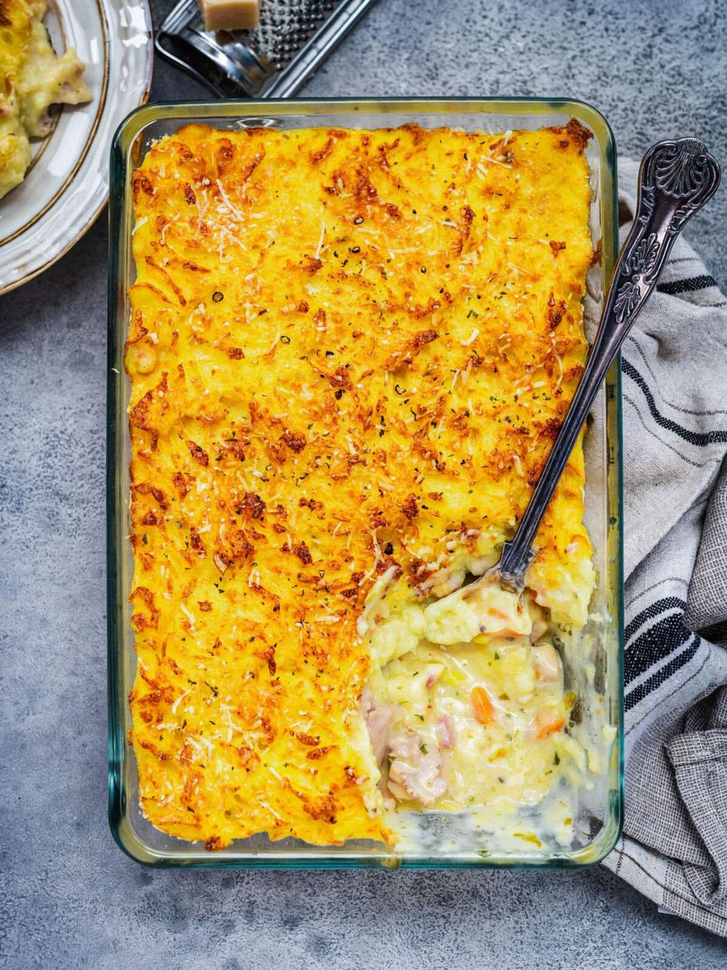 Rectangular glass dish filled with a golden-brown baked chicken shepherd's pie, topped with grated cheese. A serving spoon is in the dish, revealing a creamy filling with vegetables and chunks, next to a striped cloth napkin.