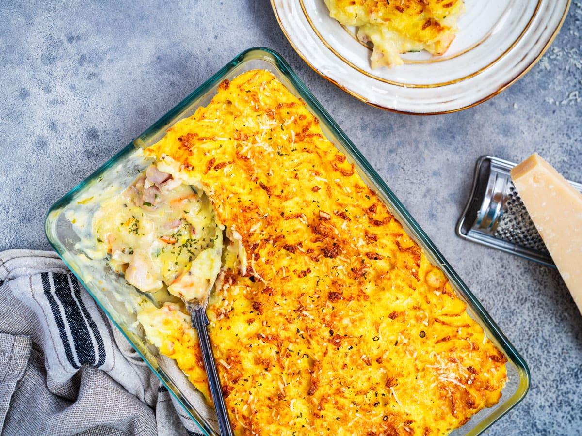A baked casserole dish filled with creamy chicken and vegetable pie topped with golden mashed potatoes. A serving has been scooped onto a plate nearby, and a cheese grater with a wedge of cheese is next to the dish.
