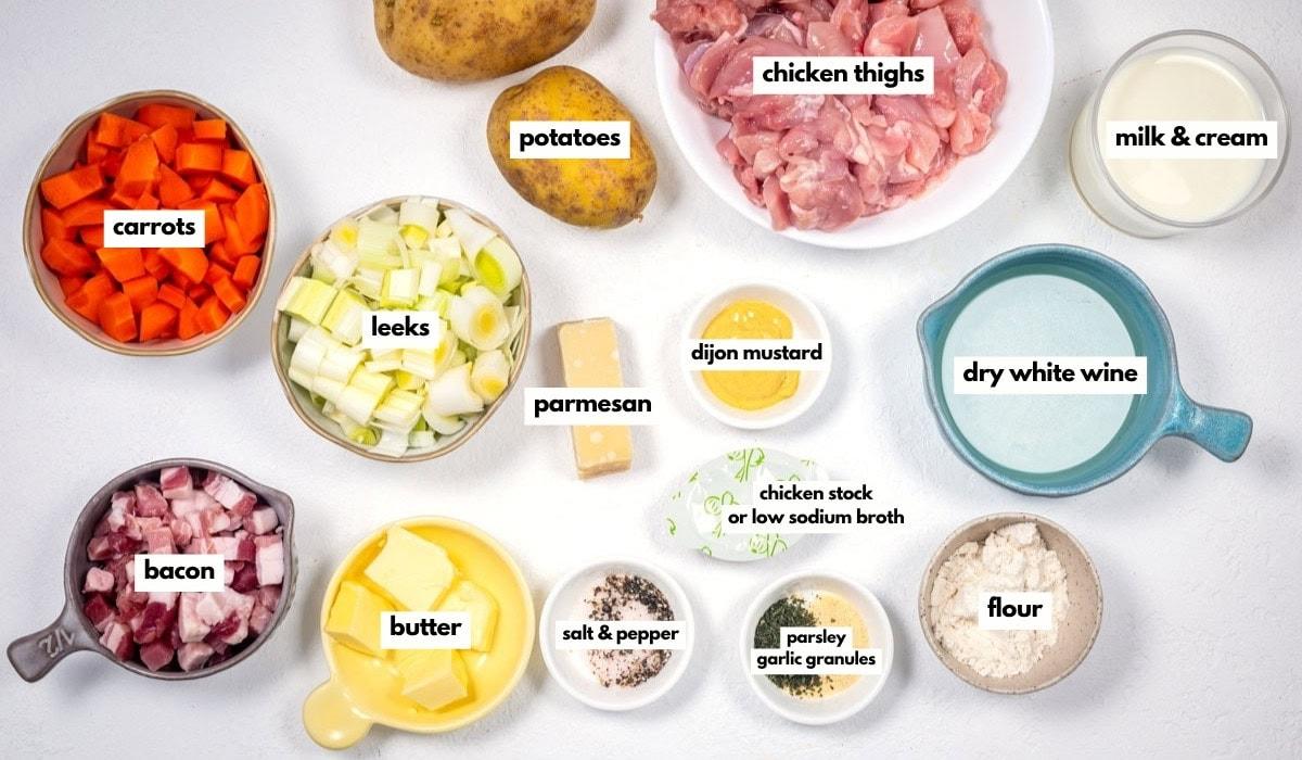 A flat lay of labeled ingredients for a shepherd's pie recipe: chicken thighs, potatoes, milk & cream, dry white wine, flour, parsley garlic granules, salt & pepper, butter, bacon, carrots, leeks, parmesan, dijon mustard, and chicken stock.