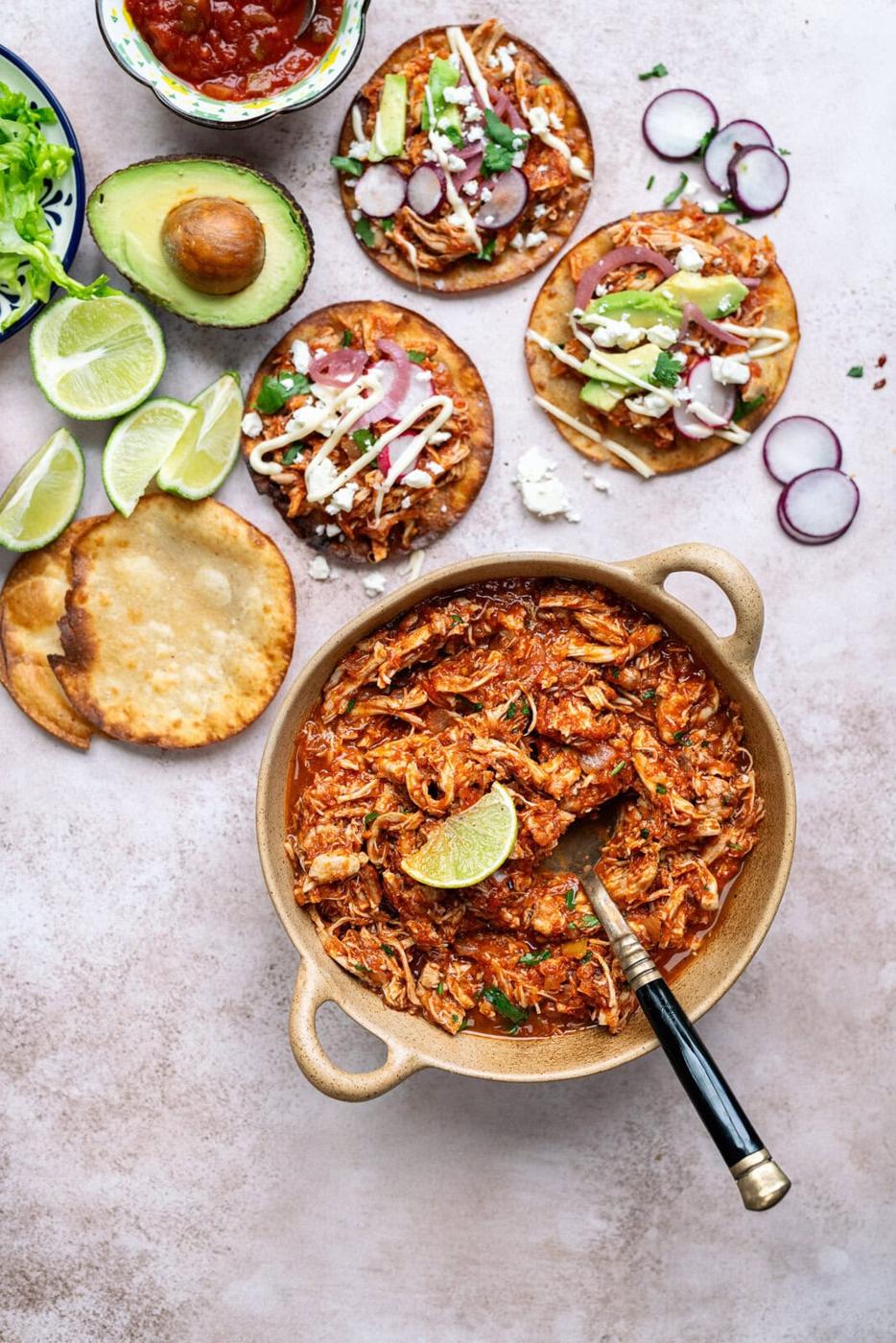 Bowl of shredded chicken tinga with tostadas on the side, avocados, lime wedges and sliced radishes