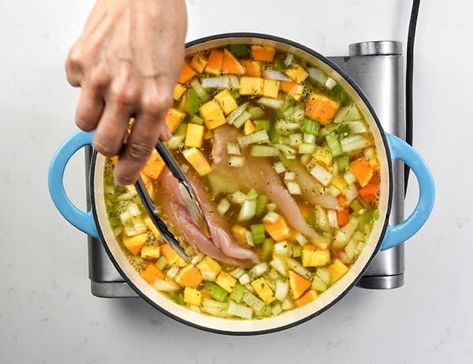A hand uses tongs to place raw chicken breasts into a pot filled with diced vegetables and broth, beginning the process of making a hearty chicken vegetable soup on the stovetop.