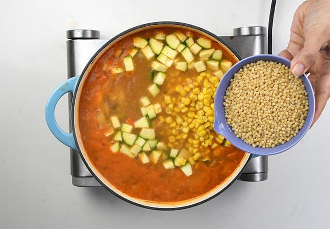 A hand holds a bowl of uncooked couscous above a pot of hearty chicken vegetable soup with zucchini, corn, and tomato broth on the stovetop, preparing to add the couscous for a filling finish.