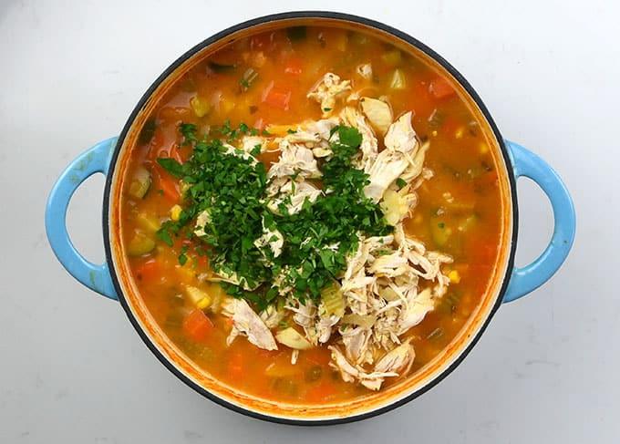 A blue pot filled with Hearty Chicken Vegetable Soup, topped with shredded chicken and chopped fresh parsley, sits on a white surface. The soup features visible pieces of vegetables like carrots and celery.