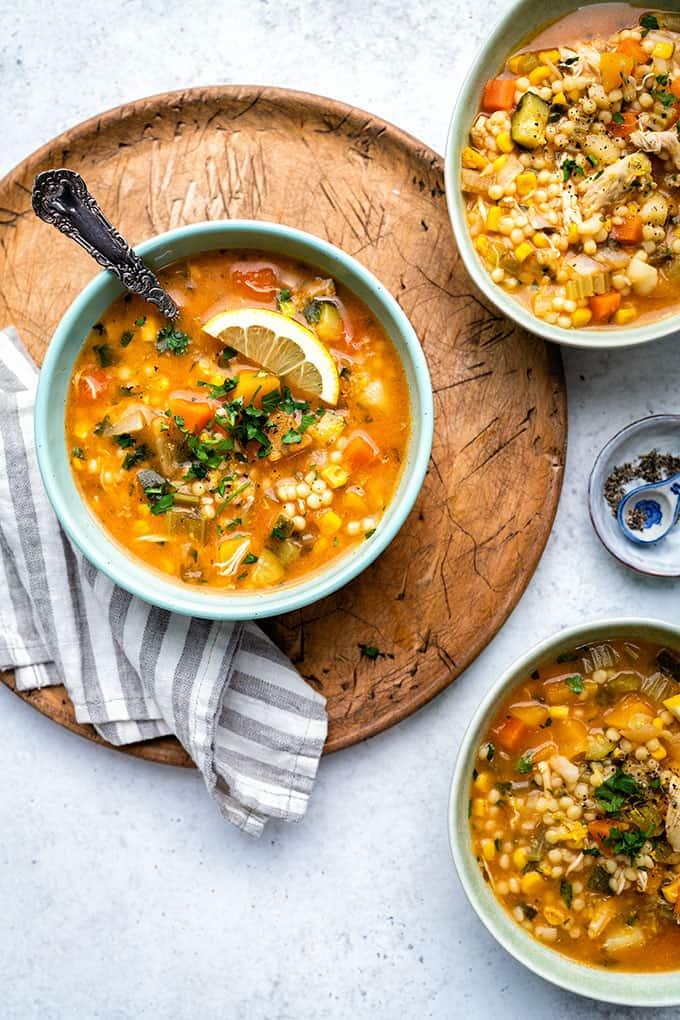 Three bowls of hearty chicken vegetable soup with barley, garnished with fresh herbs and a lemon slice, sit on a wooden board and striped napkin, with a small dish of pepper on the side.