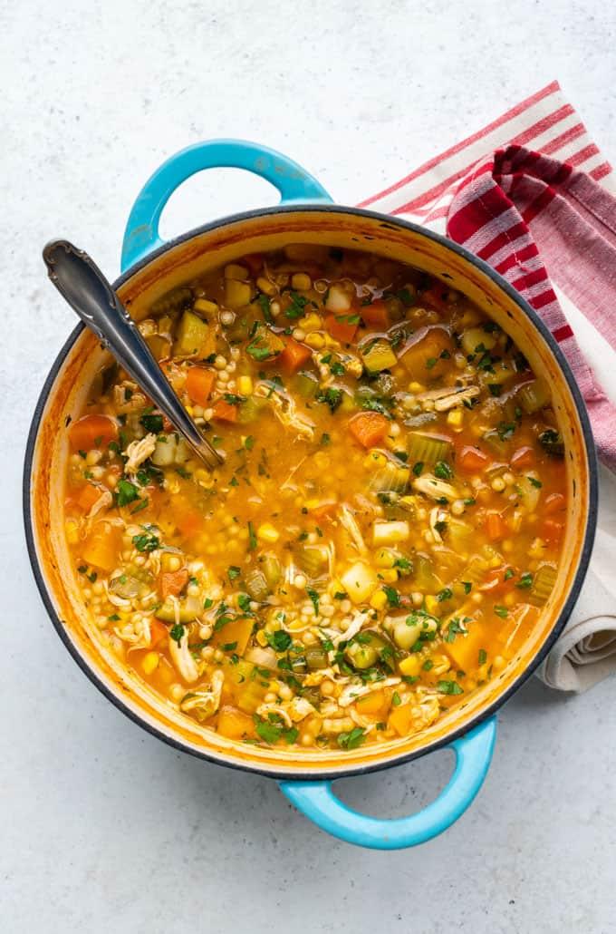 A blue pot filled with hearty chicken vegetable soup, colorful vegetables, barley, and herbs. Visible carrots, corn, and celery fill the dish. A large spoon rests inside the pot, with a red-striped towel nearby.