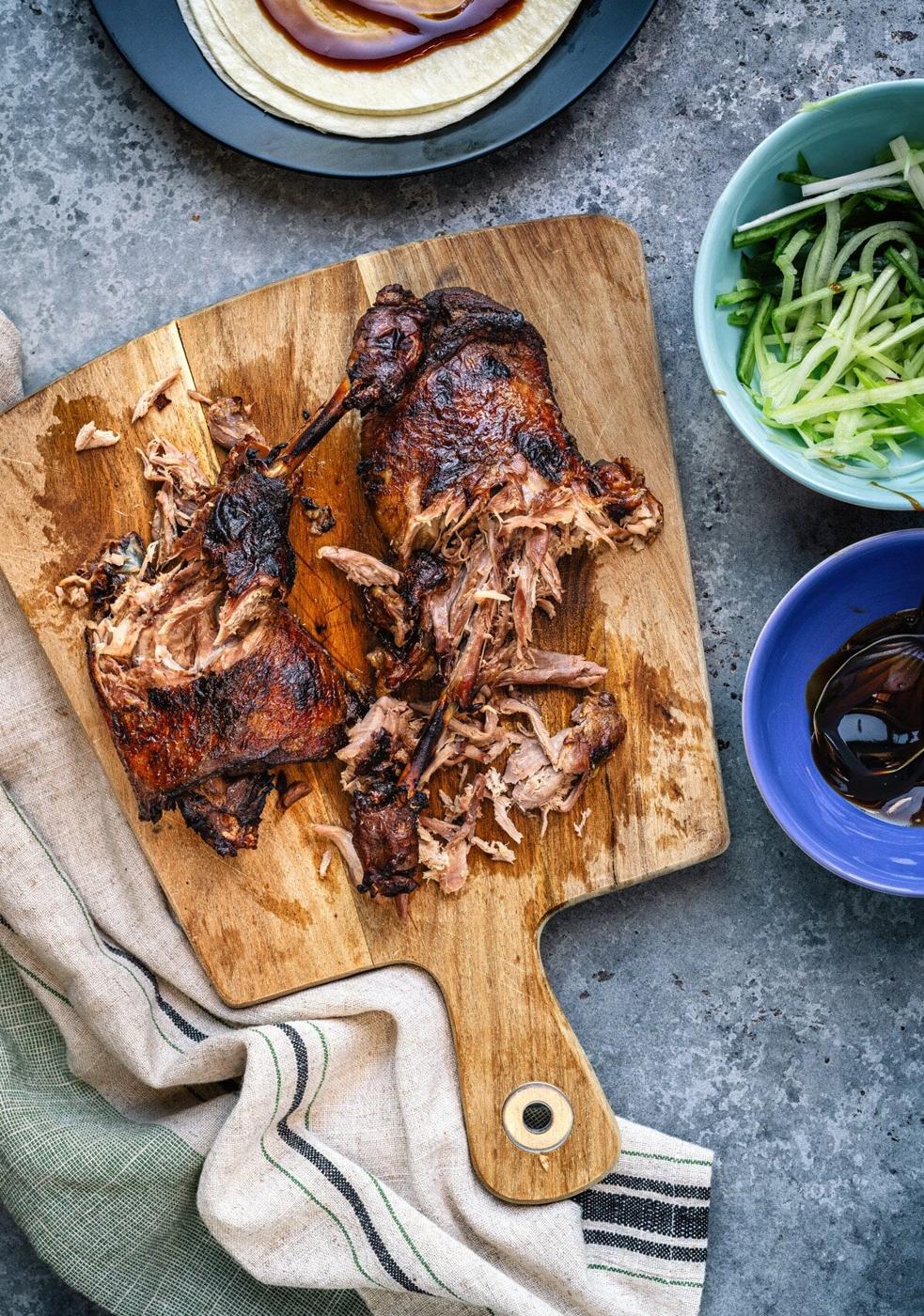 Chinese crispy duck, shredded on a wooden cutting board, is surrounded by a beige towel, cucumber and green onion strips in a bowl, and a small dish of dark sauce on a gray stone surface. Perfect for recreating at home—try it in your air fryer!.