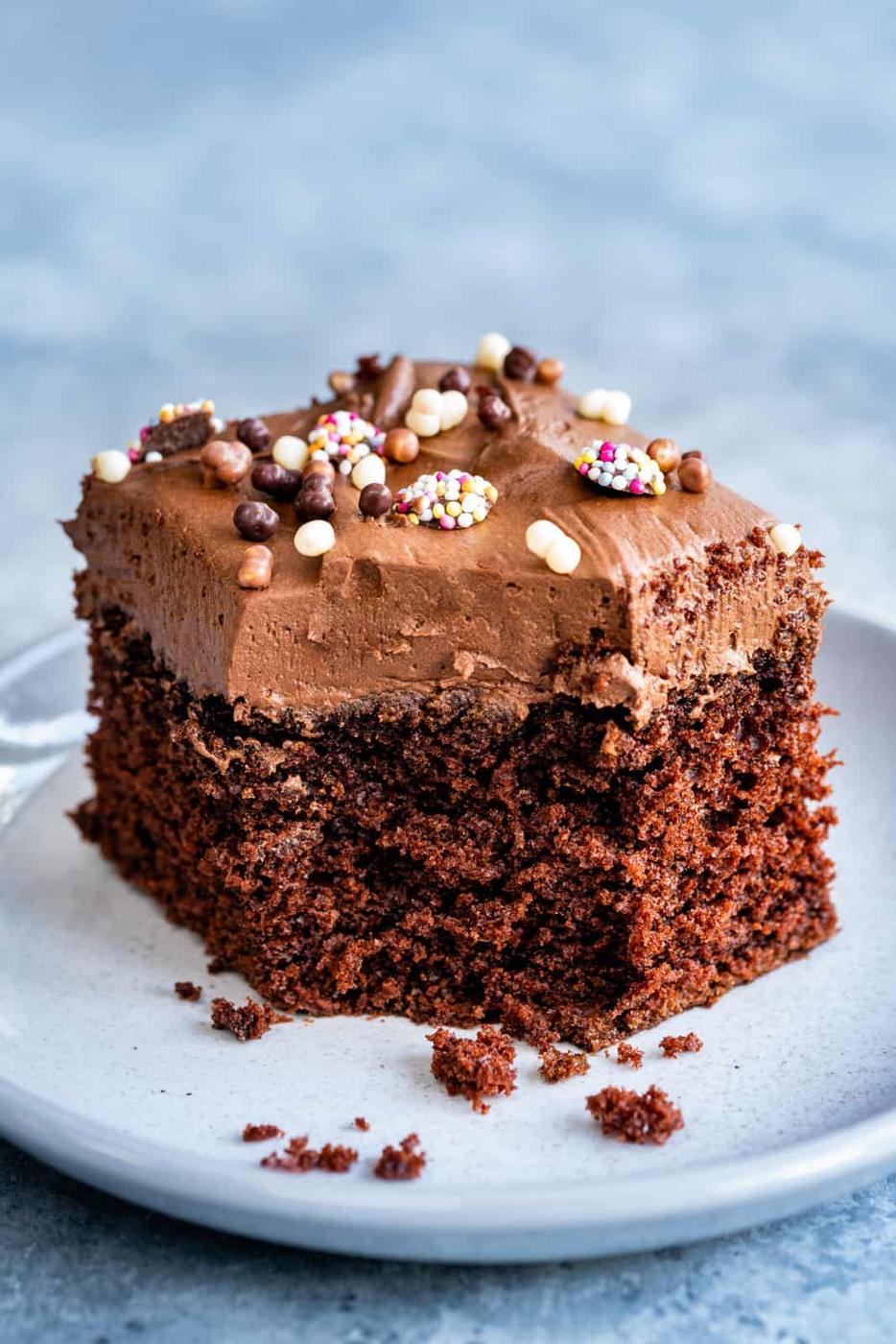 A square slice of chocolate sheet cake with chocolate frosting and colorful sprinkles sits on a white plate, with a few crumbs scattered around.