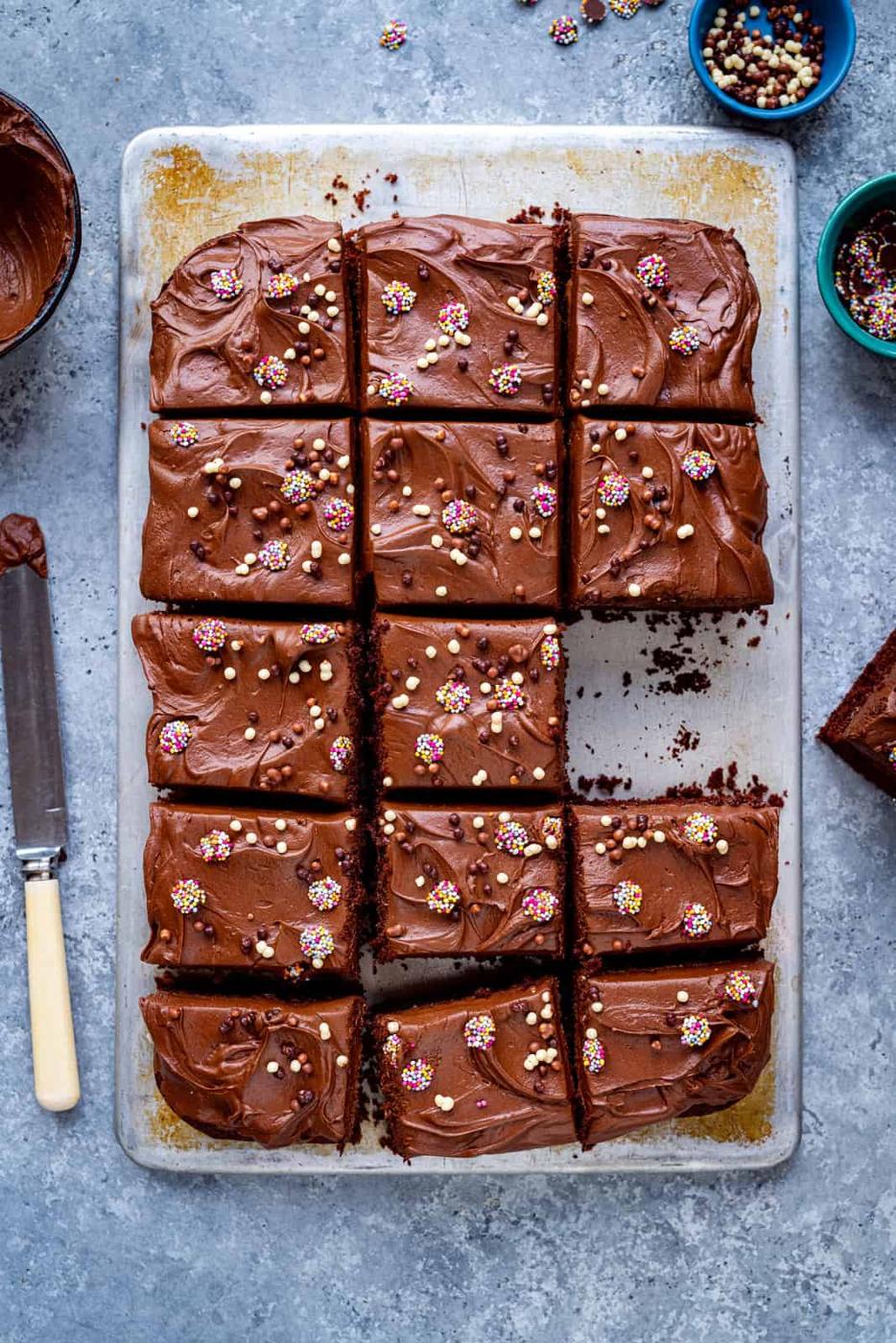 An overhead view of chocolate sheet cake (chocolate traybake) cut into neat squares, each topped with chocolate frosting and colorful sprinkles, with a few pieces missing. A knife and bowls of extra sprinkles are nearby on the gray surface.