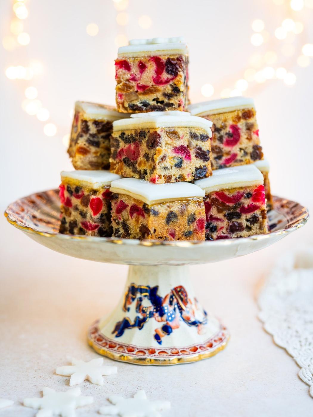 Slices of Mary Berry's fruit cake, topped with white icing, are stacked on a decorative cake stand. The colorful pieces create delightful Christmas Cake Bites, set against a festive backdrop of blurred lights.