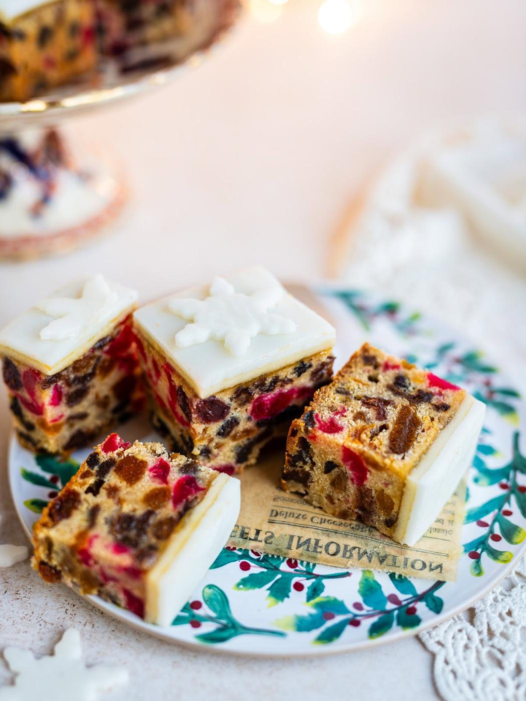 Four pieces of fruitcake topped with white icing and snowflake decorations are arranged on a festive plate. The fruitcake is filled with colorful candied fruit and nuts. A cake stand and more fruitcake are in the background.