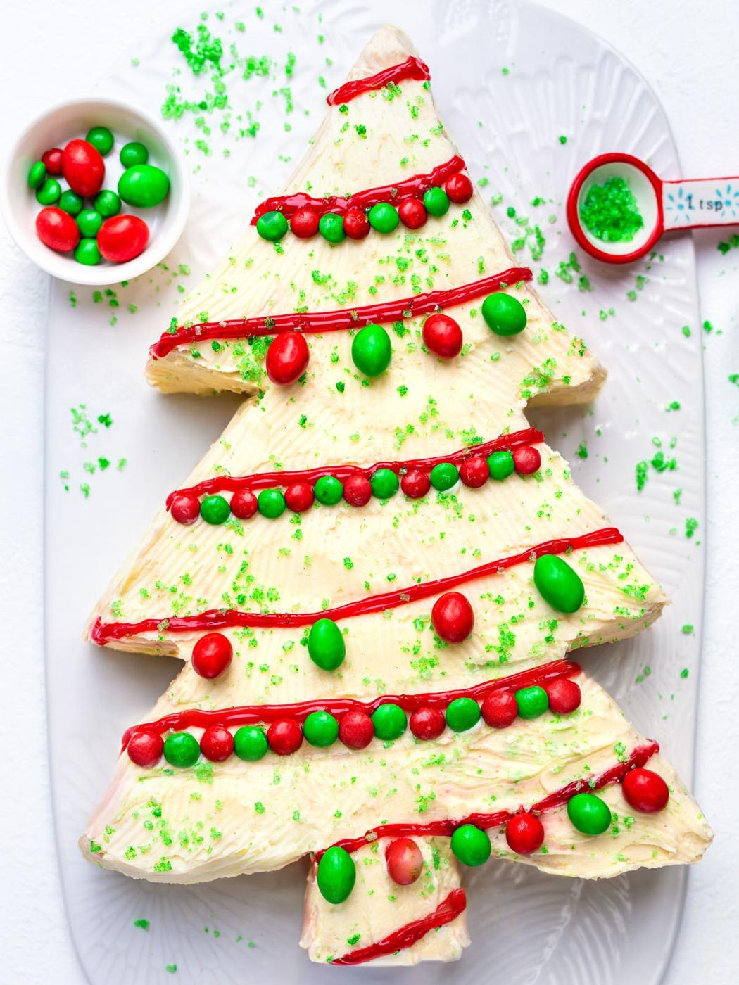 A Christmas tree-shaped cake with white frosting, decorated with red icing, green sugar sprinkles, and red and green candies, sits on a white platter. A small bowl of candies and a teaspoon of sprinkles are nearby.