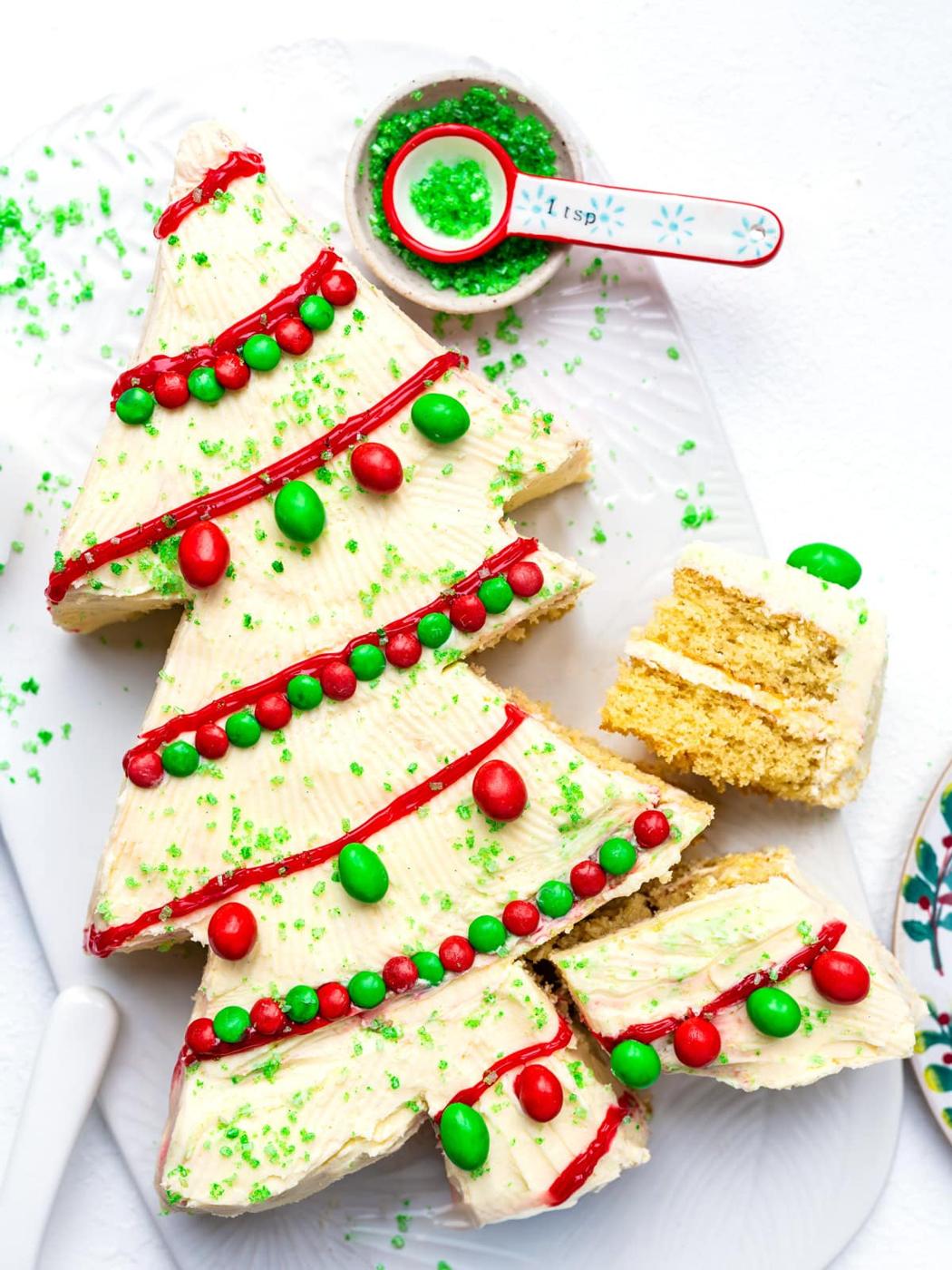 A Christmas tree-shaped cake with white frosting, red icing garlands, green and red candies as ornaments, and green sugar sprinkles. A slice is cut out, and a spoon with green sprinkles is beside the cake.