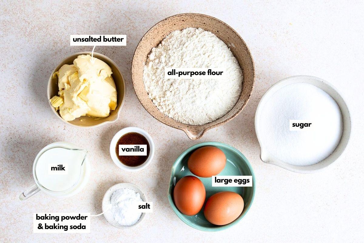 A flat lay of baking ingredients in bowls: unsalted butter, all-purpose flour, sugar, milk, vanilla, large eggs, salt, baking powder, and baking soda, each labeled with text on the image.