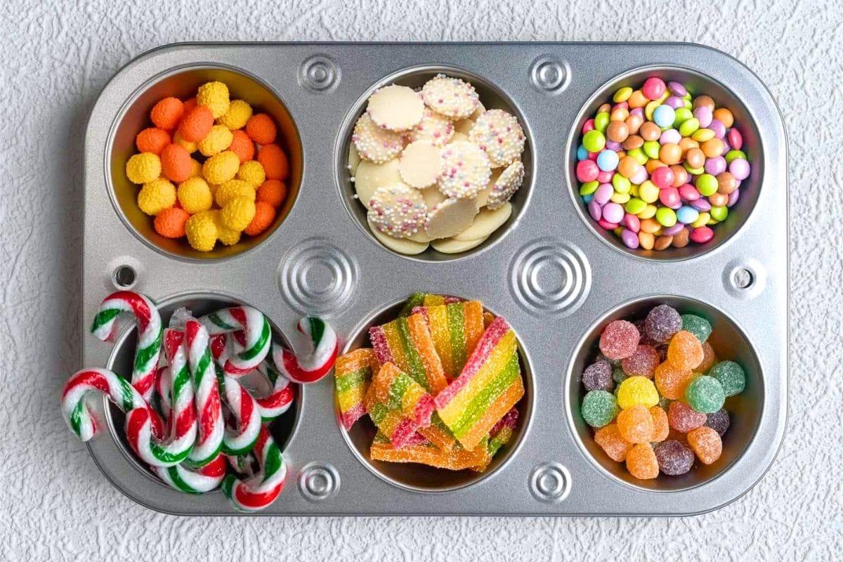 A muffin tray with six compartments filled with various colorful candies, including gummies, candy canes, chocolate buttons, and round sugar-coated sweets, placed on a textured white surface.