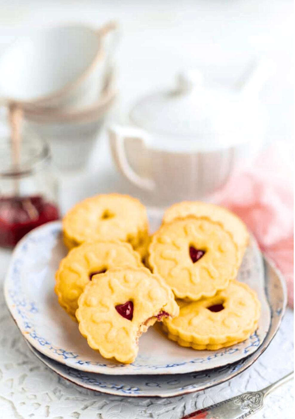A plate of five round Jammie Dodgers with scalloped edges and a red jam center, one with a bite taken out. In the background are stacked bowls, a white teapot, and a jar of jam.