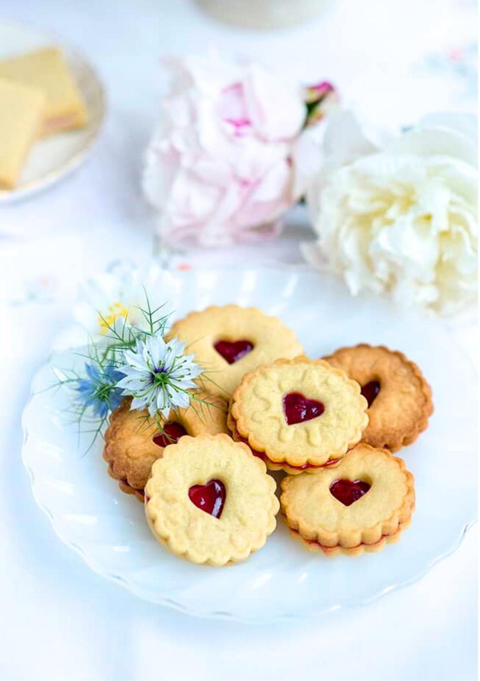 A white plate holds six round Jammie Dodgers cookies with scalloped edges and heart-shaped jam centers, garnished with small blue and white flowers. Soft-focus flowers are visible in the background.