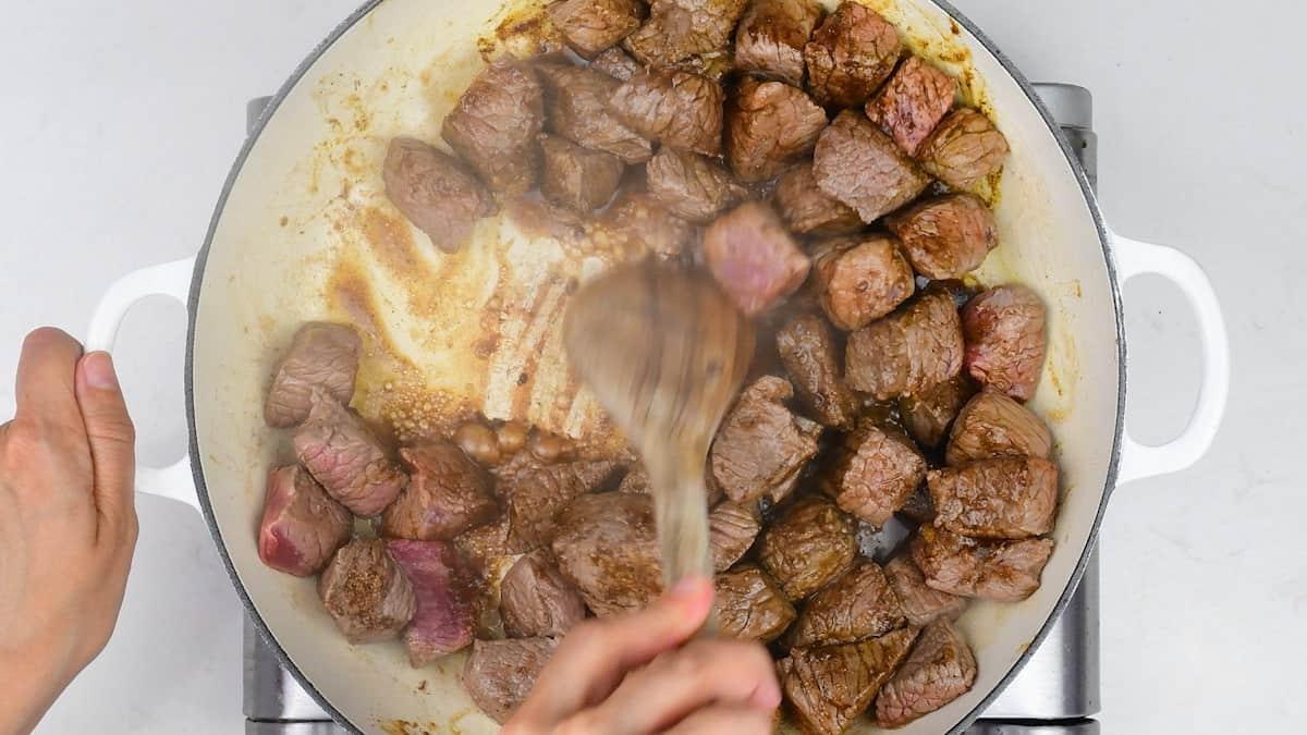 using a wooden spoon to scrape the browned bits in a casserole with seared beef cubes