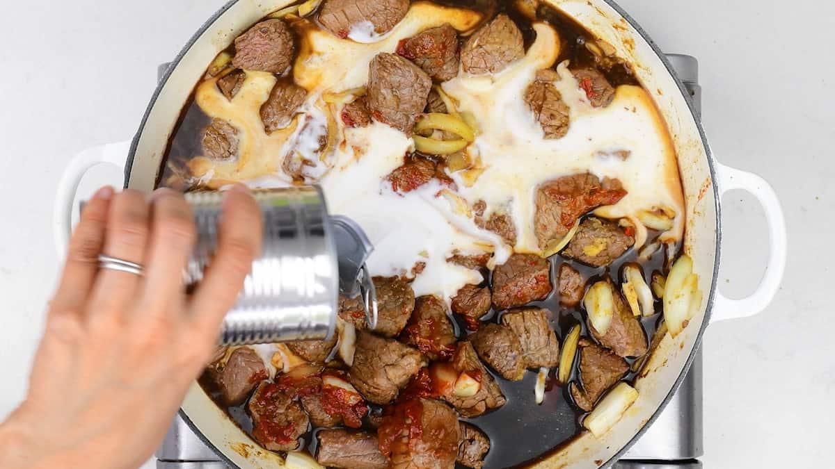 adding coconut milk to a casserole of beef curry