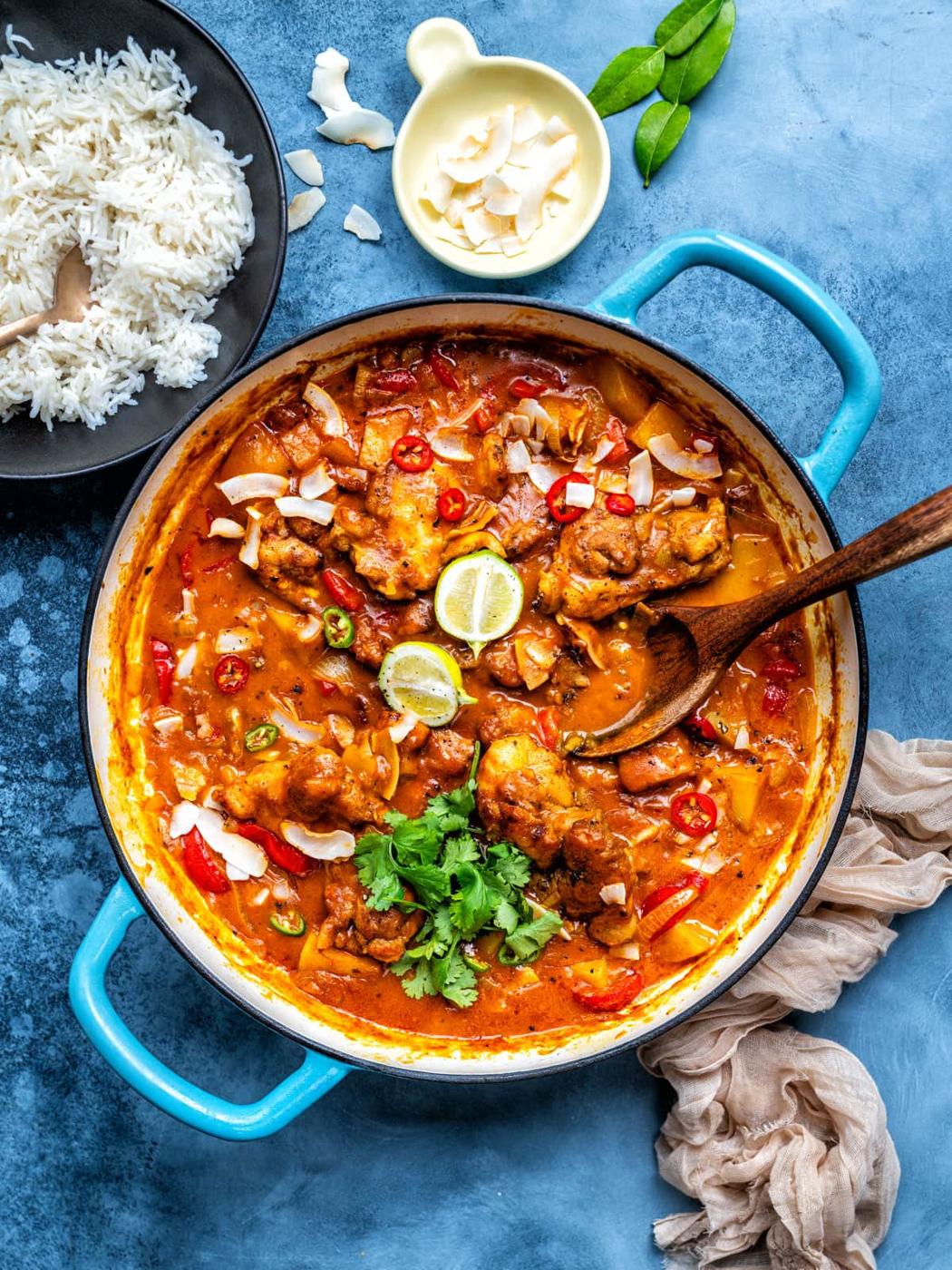 A blue pot filled with a vibrant chicken curry garnished with lime slices, cilantro, and chili, with a wooden spoon inside. Nearby are a bowl of white rice and a small dish of coconut flakes on a blue surface.