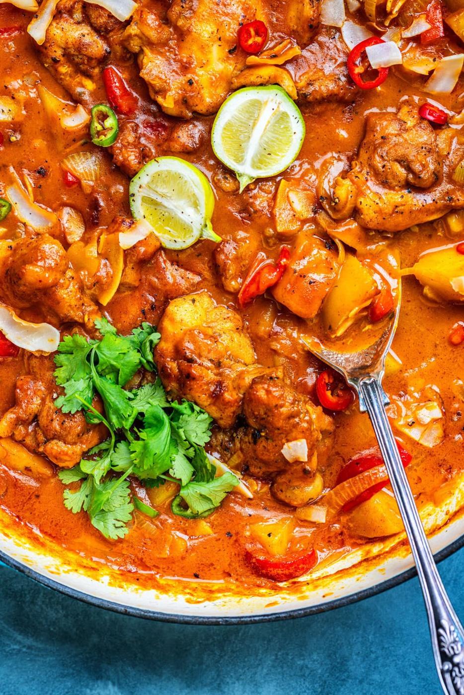 A close-up of a vibrant chicken curry with pieces of chicken, bell peppers, sliced lime, red chilies, fresh cilantro, and a rich orange sauce, served in a large pan with a spoon.