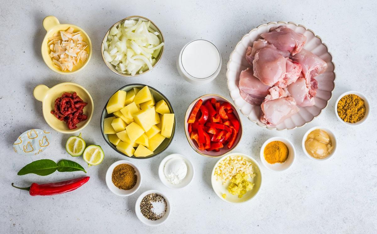 A variety of ingredients arranged neatly on a white surface, including raw chicken, chopped onions, potatoes, red bell pepper, spices, ginger, garlic, sliced chili, lime, coconut milk, and tomato paste in bowls and cups.