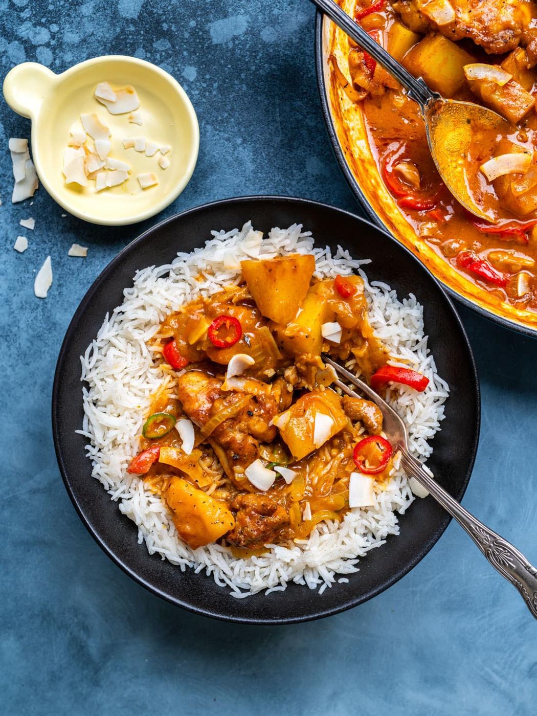 A black bowl of white rice topped with coconut chicken curry, garnished with red chili and coconut flakes. A fork rests in the bowl. A pot of curry and a small dish of coconut flakes are nearby on a blue surface.